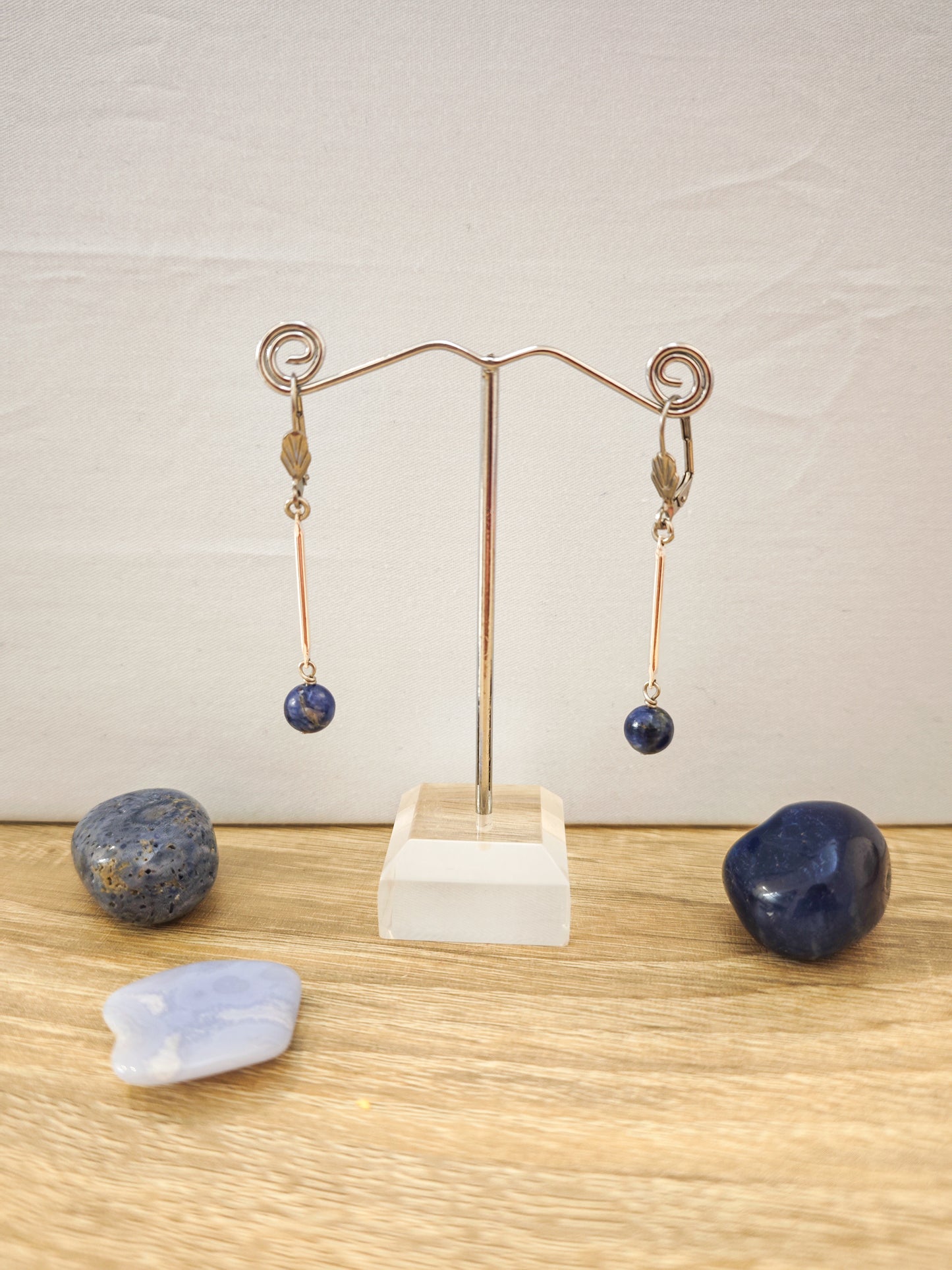 Boucles d’oreilles fines dorées avec pierres naturelles de sodalite bleue sur présentoir métallique, ambiance minimaliste et artisanale.