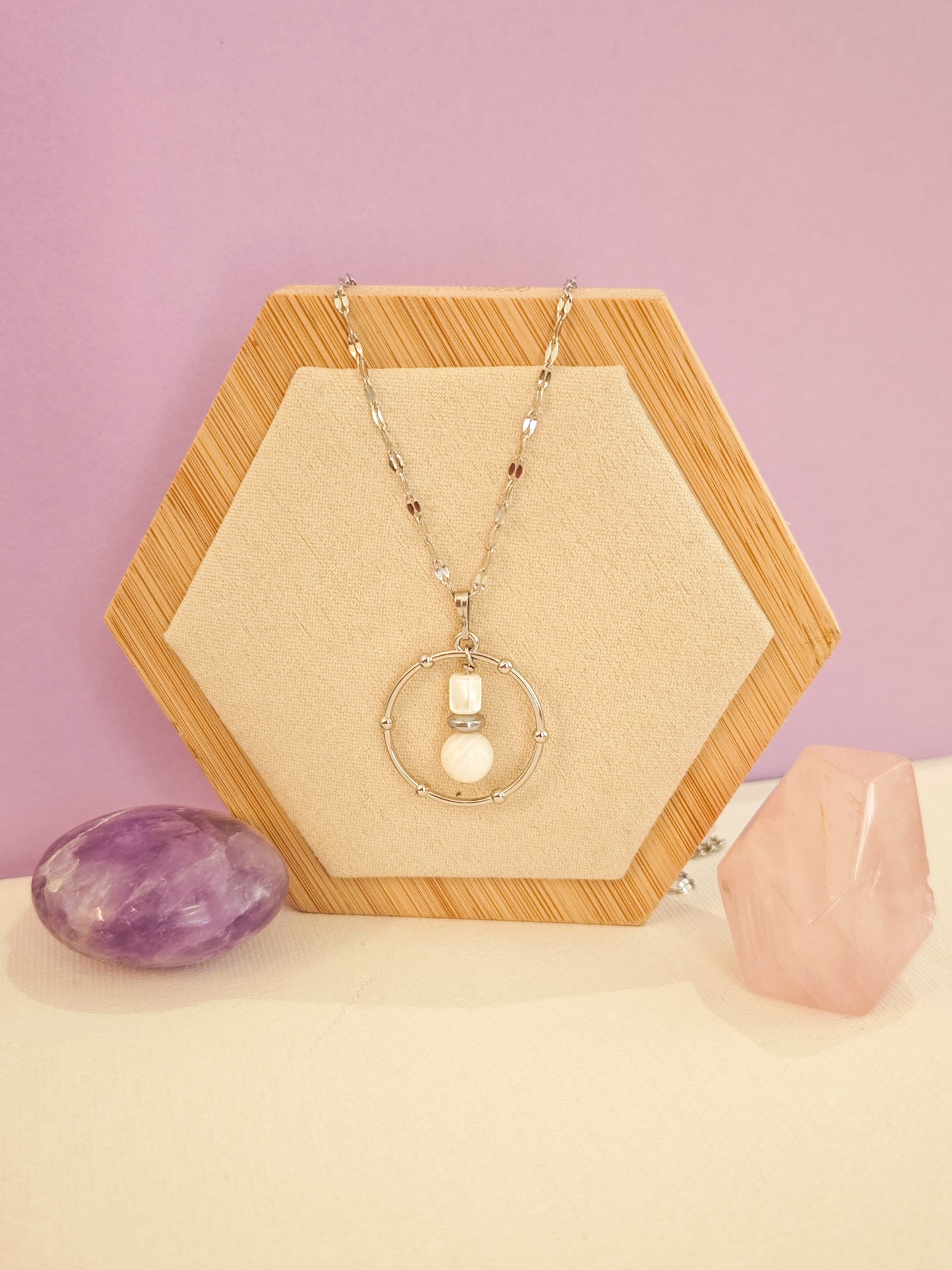 Collier artisanal en acier inoxydable avec pendentif cercle, nacre blanche et hématite, création Julie Gassmann sur fond beige et rose.