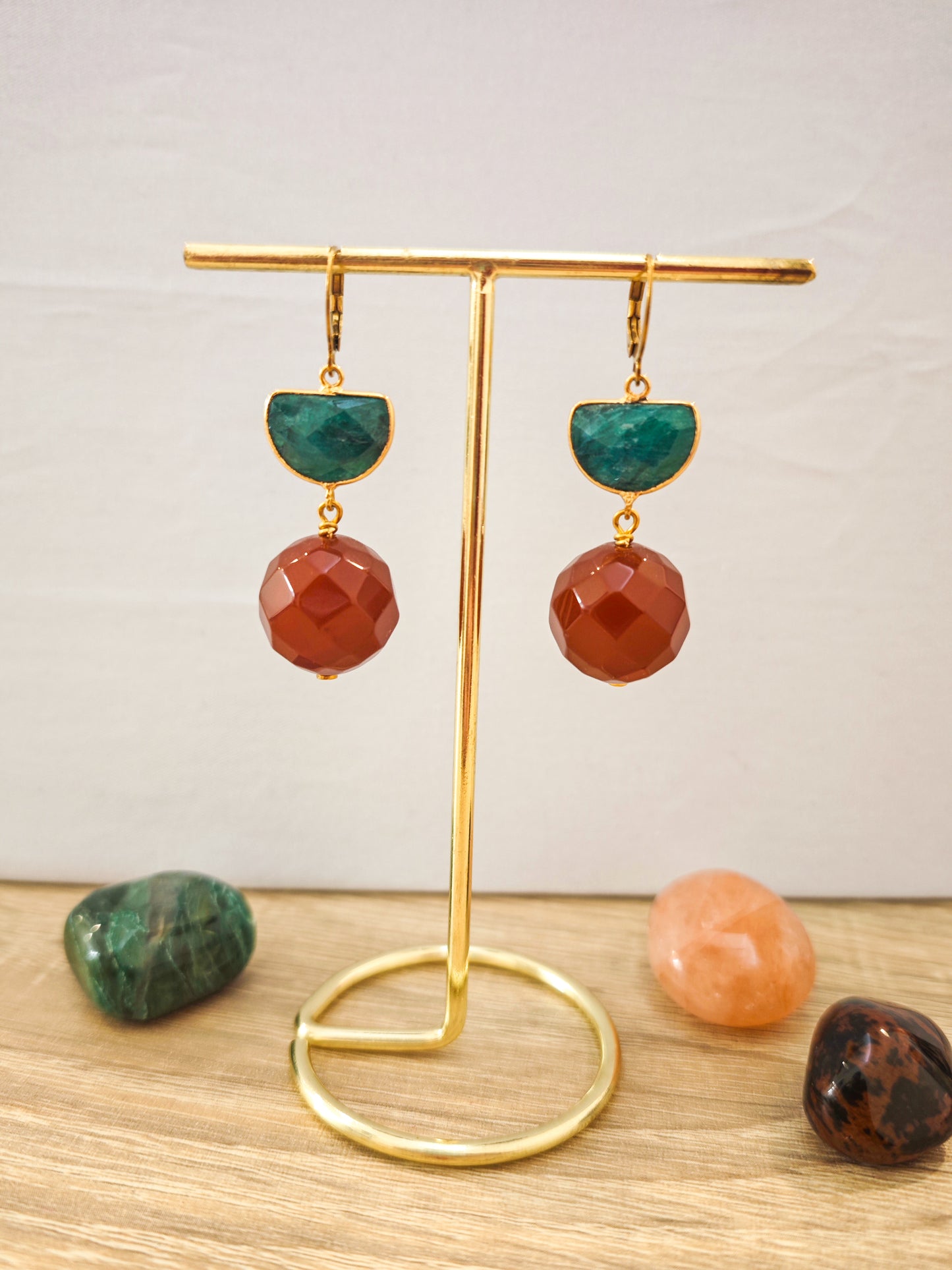 Boucles d’oreilles artisanales en émeraude verte et cornaline facettée orange sur acier inoxydable doré présentées sur un support doré.