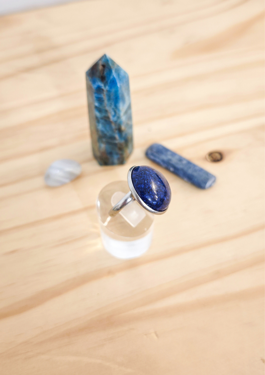 Bague artisanale en lapis-lazuli bleu marine montée sur acier inoxydable, posée sur un support transparent devant des pierres bleues sur une table en bois clair.