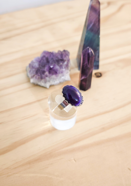 Bague artisanale en agate violette montée sur acier inoxydable, posée sur un support transparent devant des cristaux violets sur une table en bois clair.
