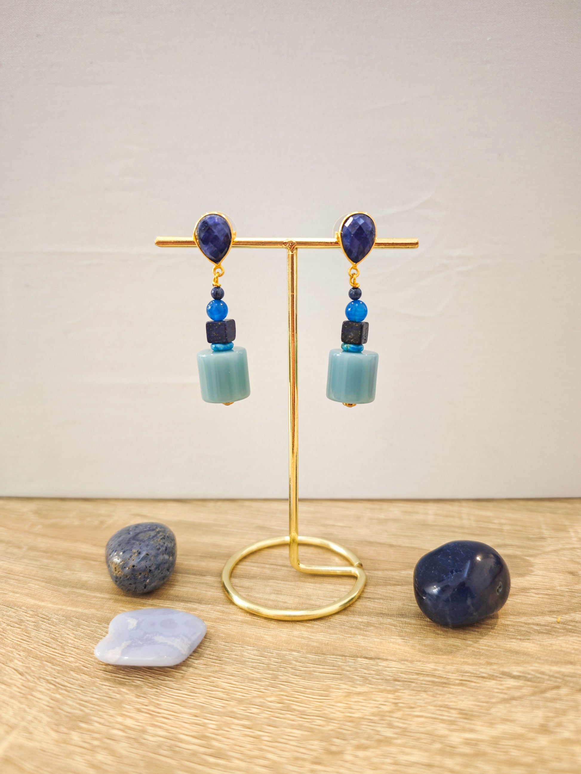 Boucles d’oreilles artisanales dorées avec lapis lazuli et perles bleues turquoise sur présentoir doré, ambiance lumineuse et élégante.