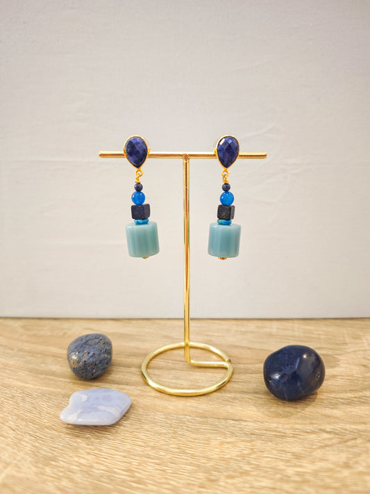 Boucles d’oreilles artisanales dorées avec lapis lazuli et perles bleues turquoise sur présentoir doré, ambiance lumineuse et élégante.