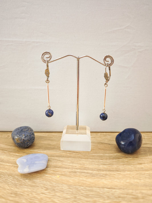 Boucles d’oreilles fines dorées avec pierres naturelles de sodalite bleue sur présentoir métallique, ambiance minimaliste et artisanale.