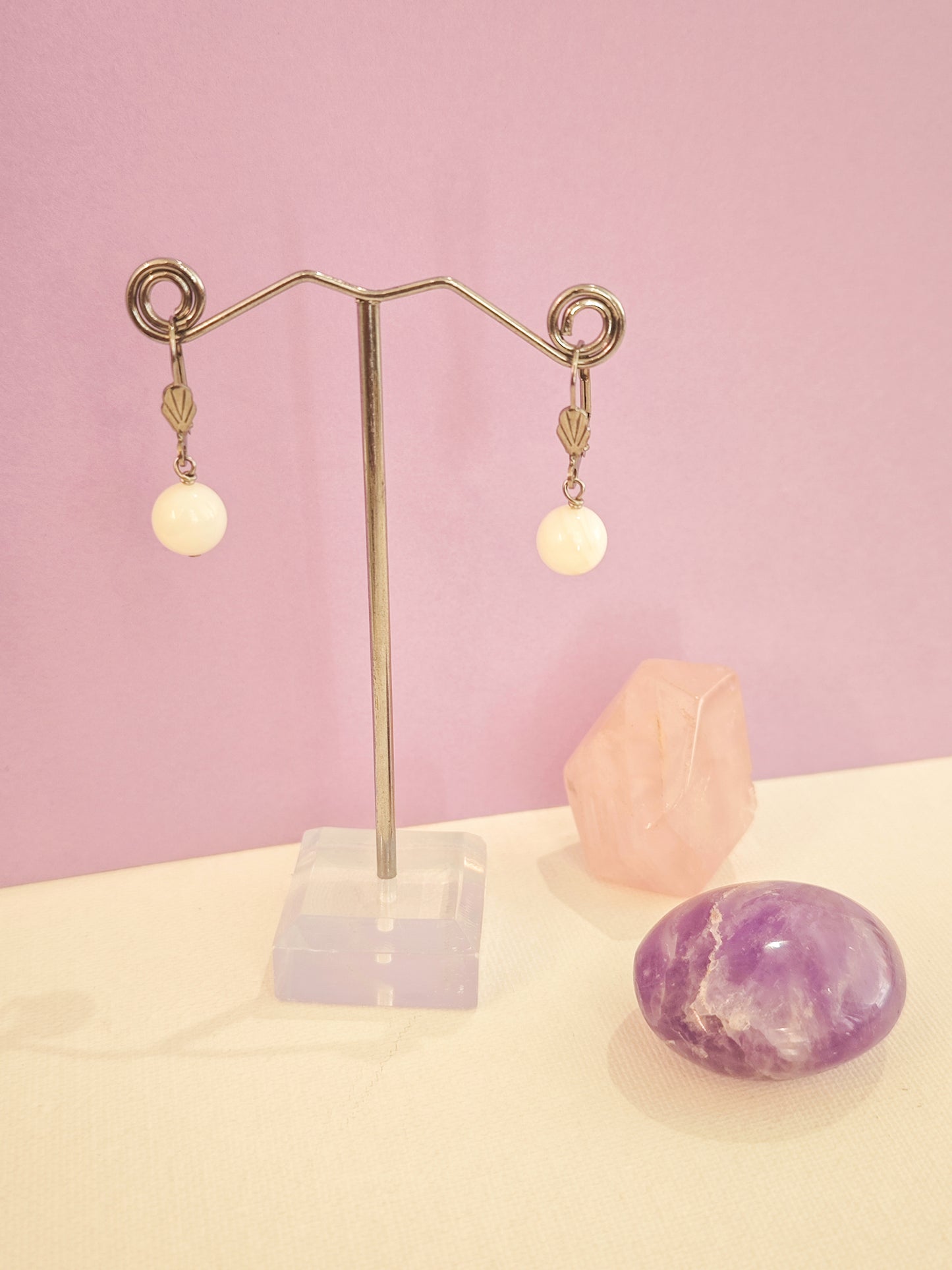 Boucles d’oreilles artisanales en acier inoxydable avec perles de nacre blanche, création Julie Gassmann sur fond rose et beige.