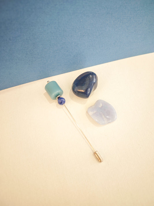 Fibule artisanale en acier avec pierres naturelles sodalite et hématite et acétate bleu, création unique Julie Gassmann sur fond beige et bleu.