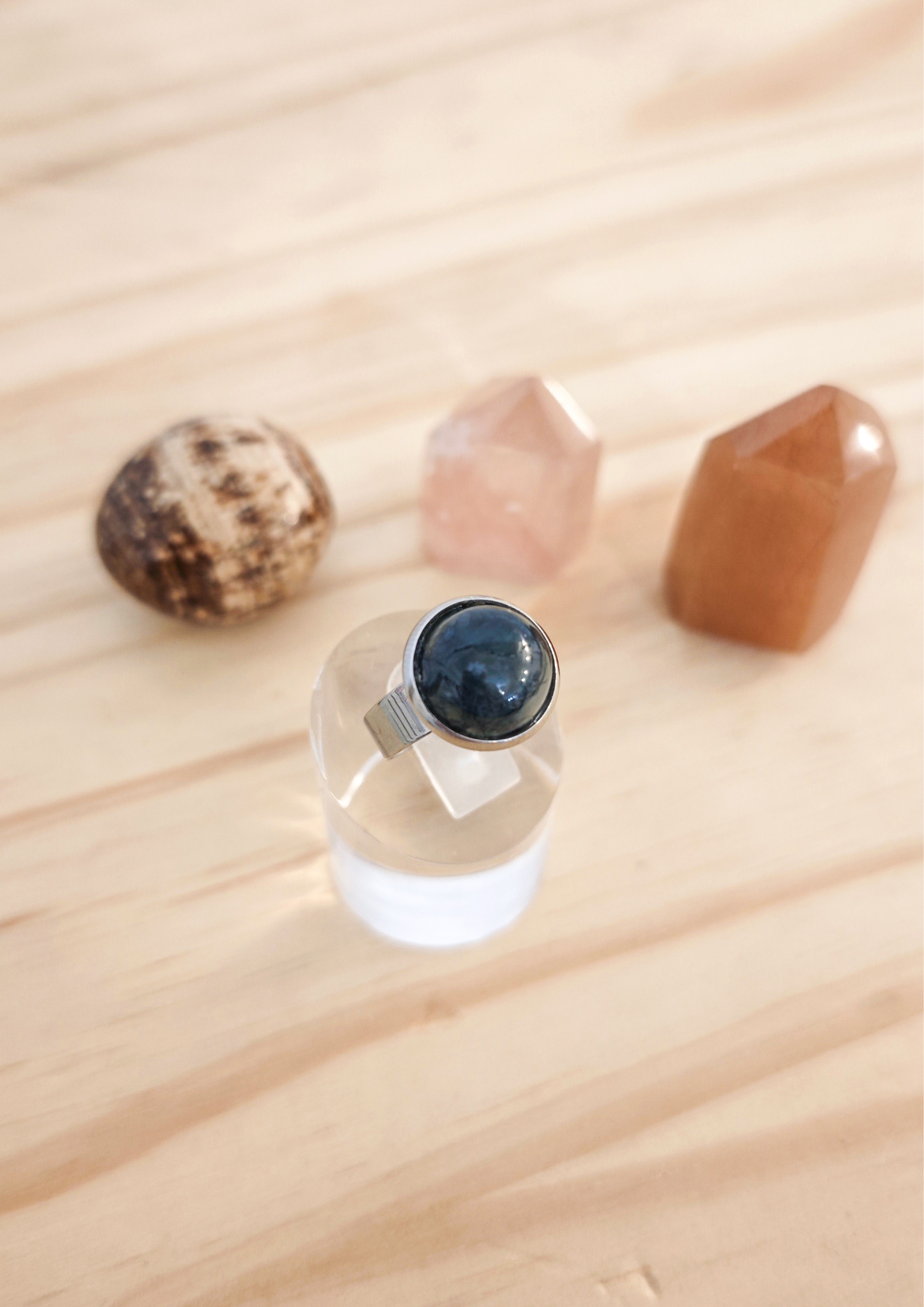 Bague ajustable en agate mousse verte montée sur acier inoxydable, posée sur un support transparent devant des pierres naturelles sur une table en bois clair.