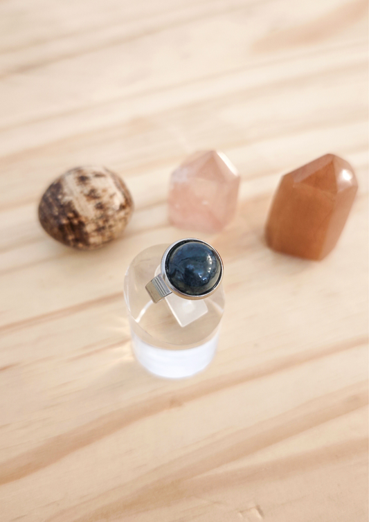 Bague ajustable en agate mousse verte montée sur acier inoxydable, posée sur un support transparent devant des pierres naturelles sur une table en bois clair.