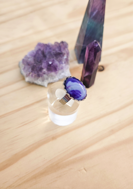 Bague artisanale en agate violette montée sur acier inoxydable, posée sur un support transparent devant des cristaux violets sur une table en bois clair.
