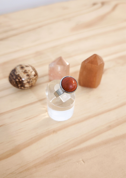 Bague ajustable en jaspe rouge montée sur acier inoxydable, posée sur un support transparent devant des pierres naturelles sur une table en bois clair.