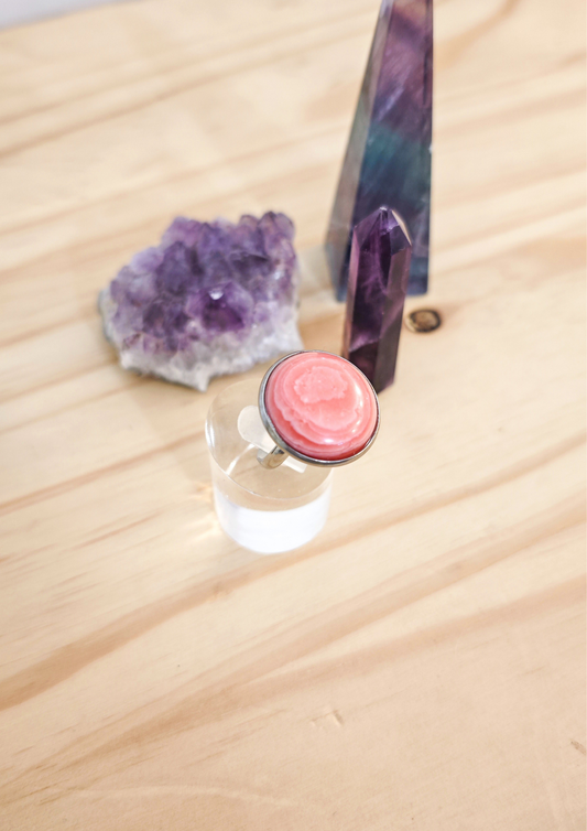 Bague artisanale en rhodochrosite rose montée sur acier inoxydable, posée sur un support transparent devant des cristaux violets sur une table en bois clair.